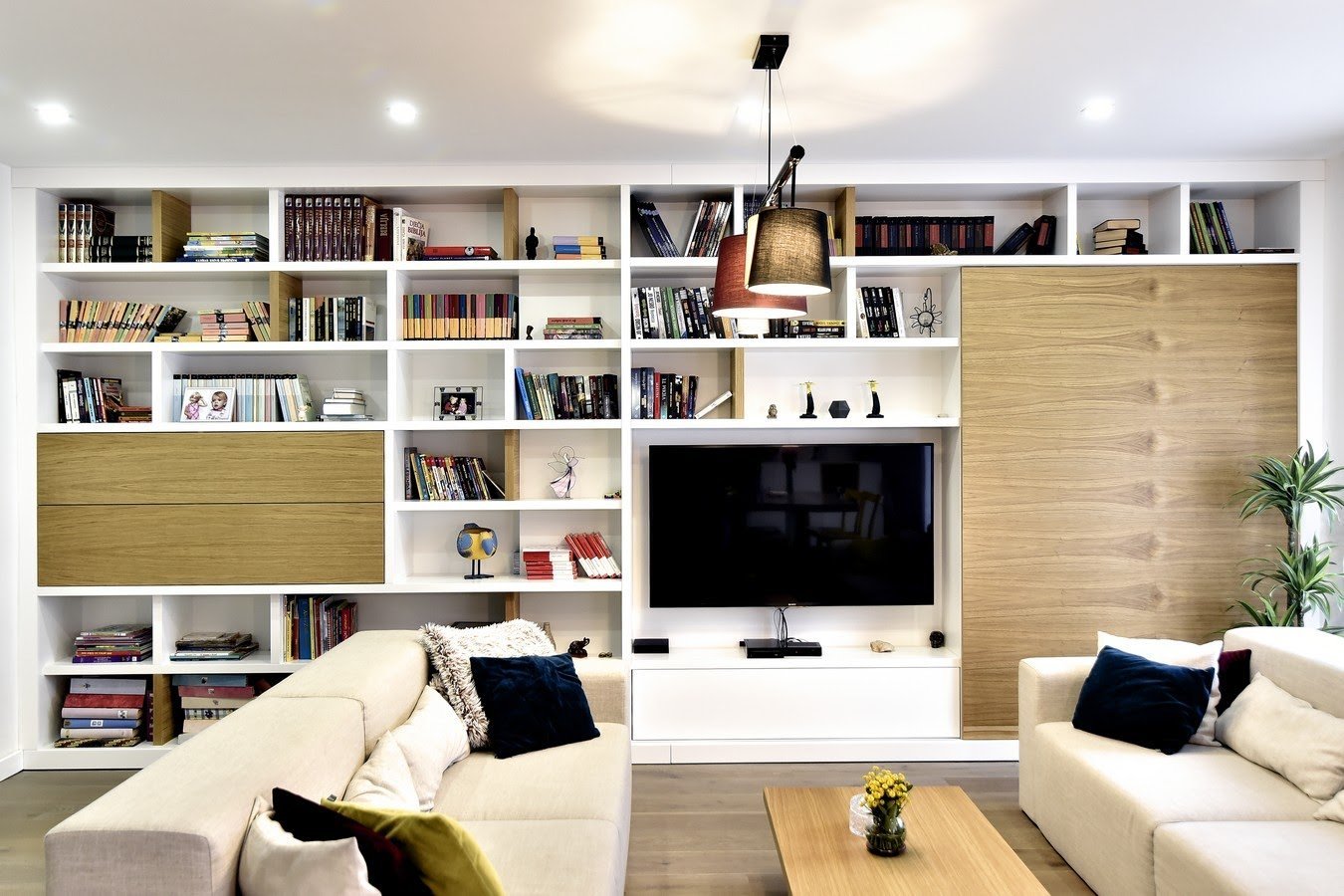 Modern living room with white built-in shelving, TV entertainment center, wooden accents, and cream-colored sofas with black pillows